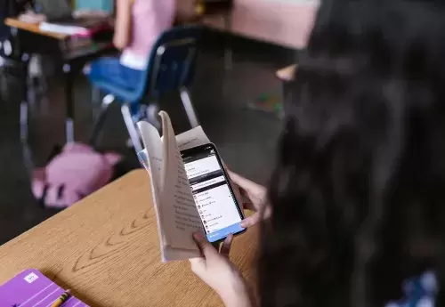youth looks at phone hidden by book at school desk