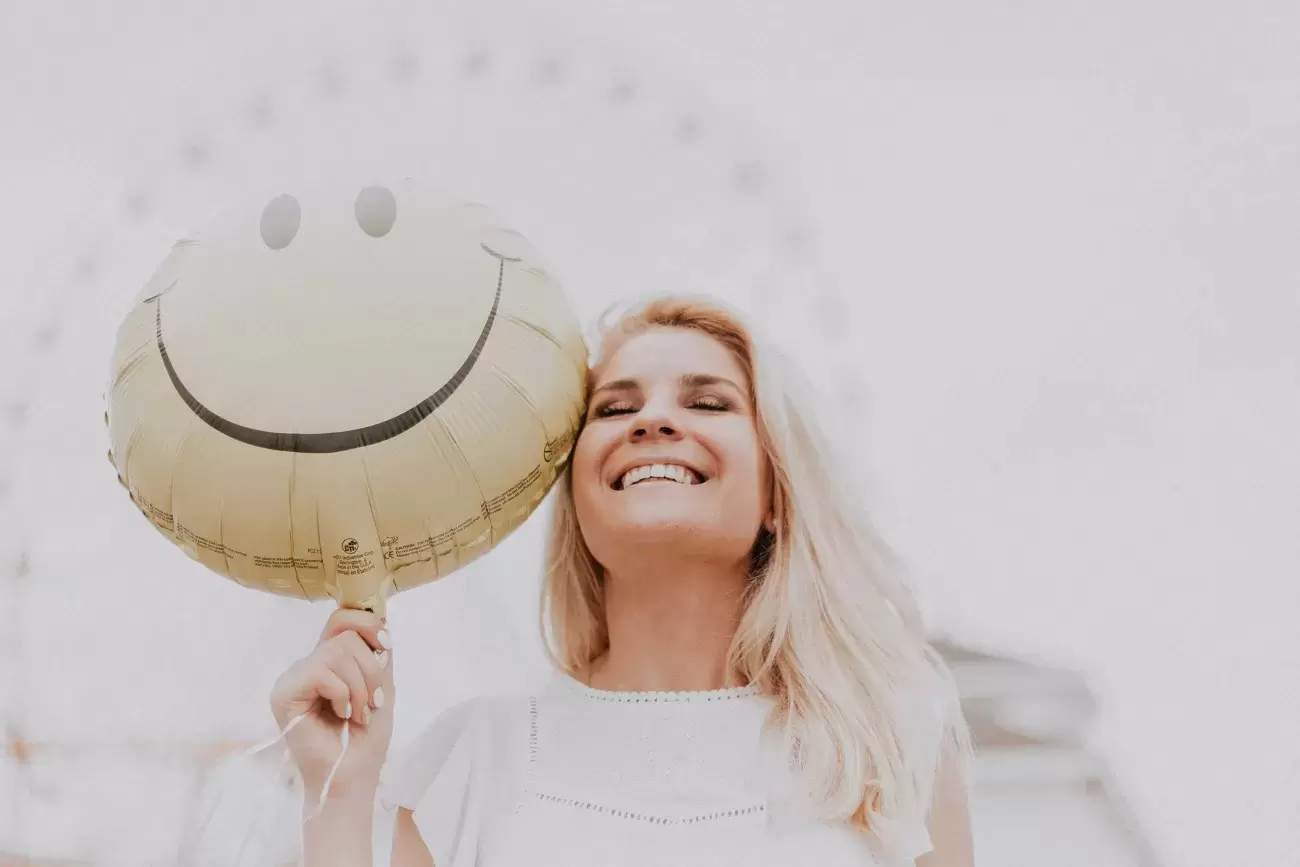 smiling person holds a yellow, happy face balloon in front of a ferris wheel